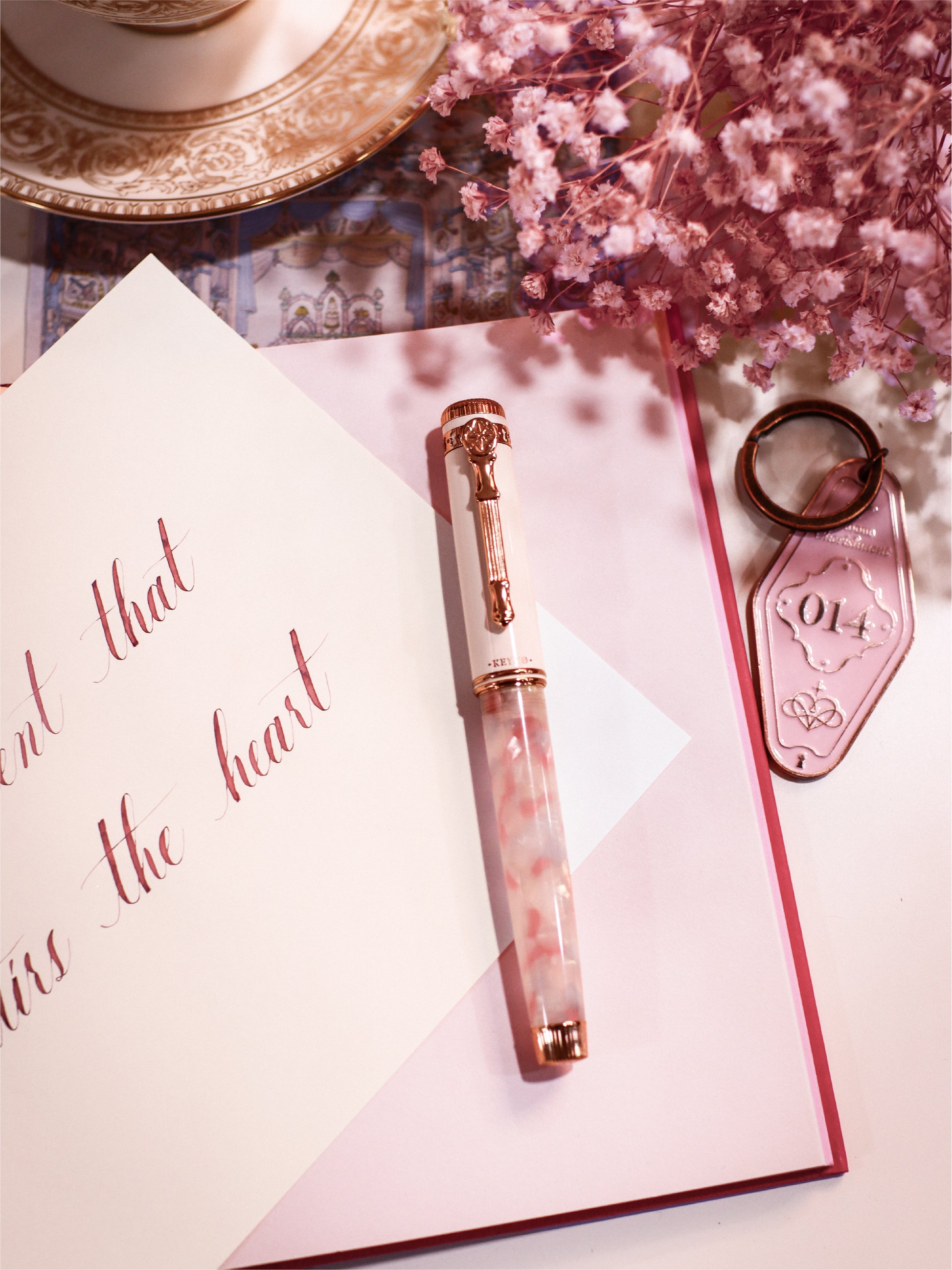 Rose gold pen on a pink notebook with a cup, saucer, and dried flowers in the background