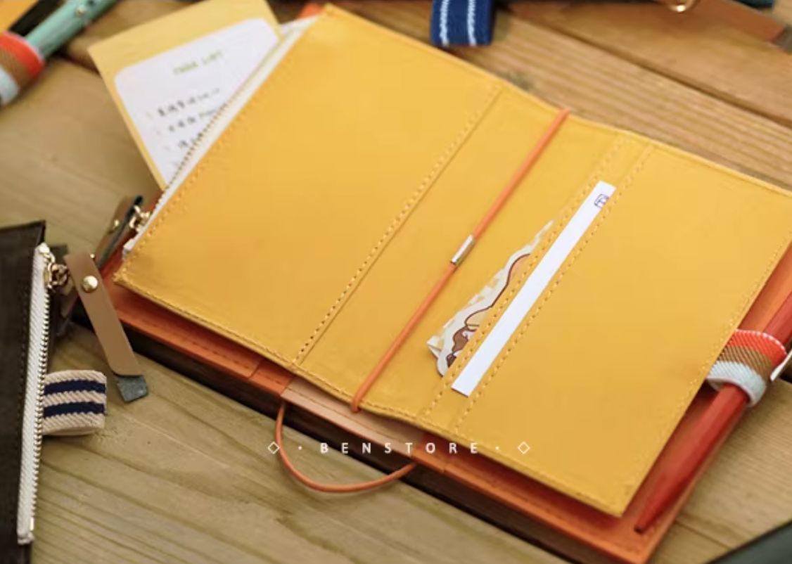 Yellow notebook with a pen holder on a wooden surface, featuring the brand 'BENSTORE'.