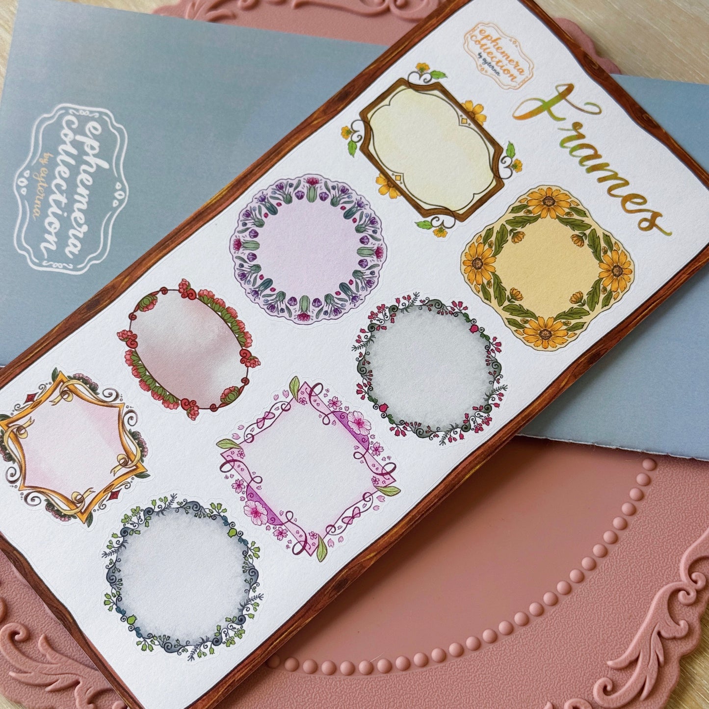 A sheet of decorative frame stickers displayed diagonally on a pink textured surface, with a blue notebook in the background. The sheet features six different floral and ornate frame designs in pastel colors with green leaves and yellow sunflower accents.