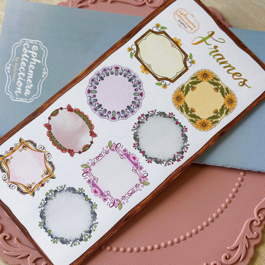 A sheet of decorative frame stickers displayed diagonally on a pink textured surface, with a blue notebook in the background. The sheet features six different floral and ornate frame designs in pastel colors with green leaves and yellow sunflower accents.