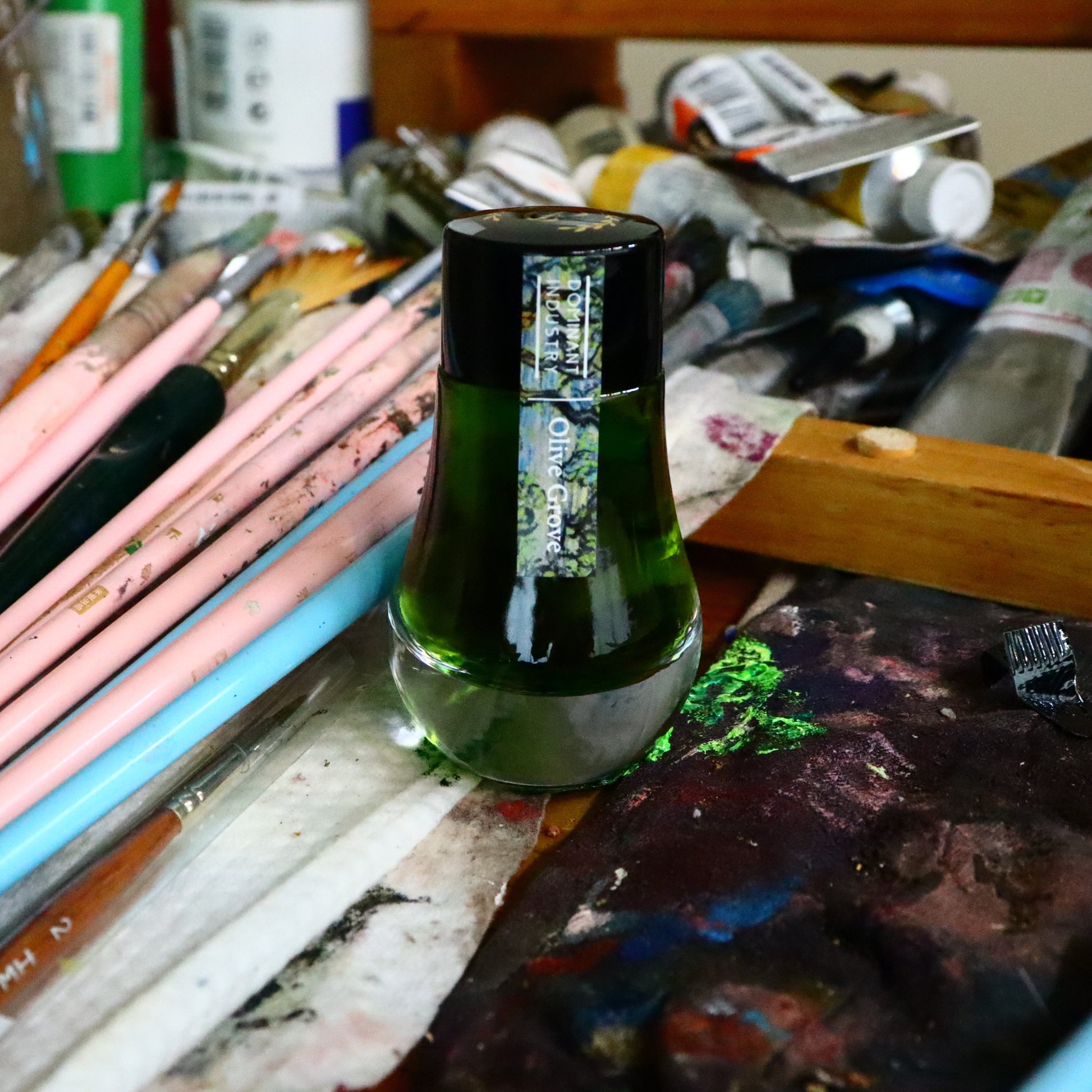 Olive Grove ink bottle on a paint palette with art supplies in the background