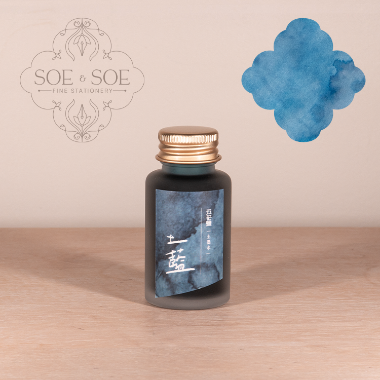 Bottle of ink with blue label and gold cap on a wooden surface, featuring SOE & SOE logo.