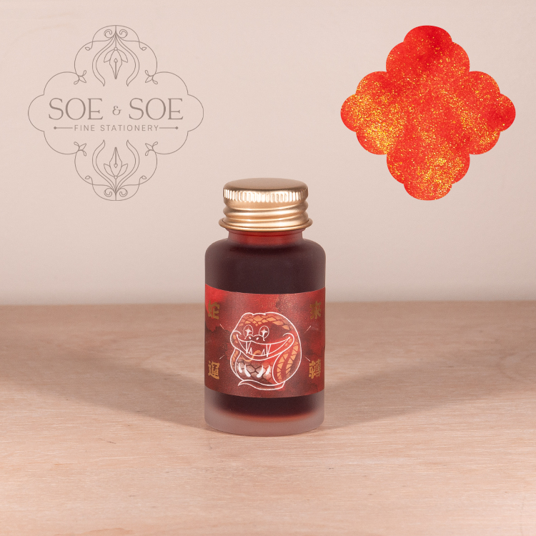 Bottle of red ink with gold cap on a wooden surface, featuring SOE & SOE branding.