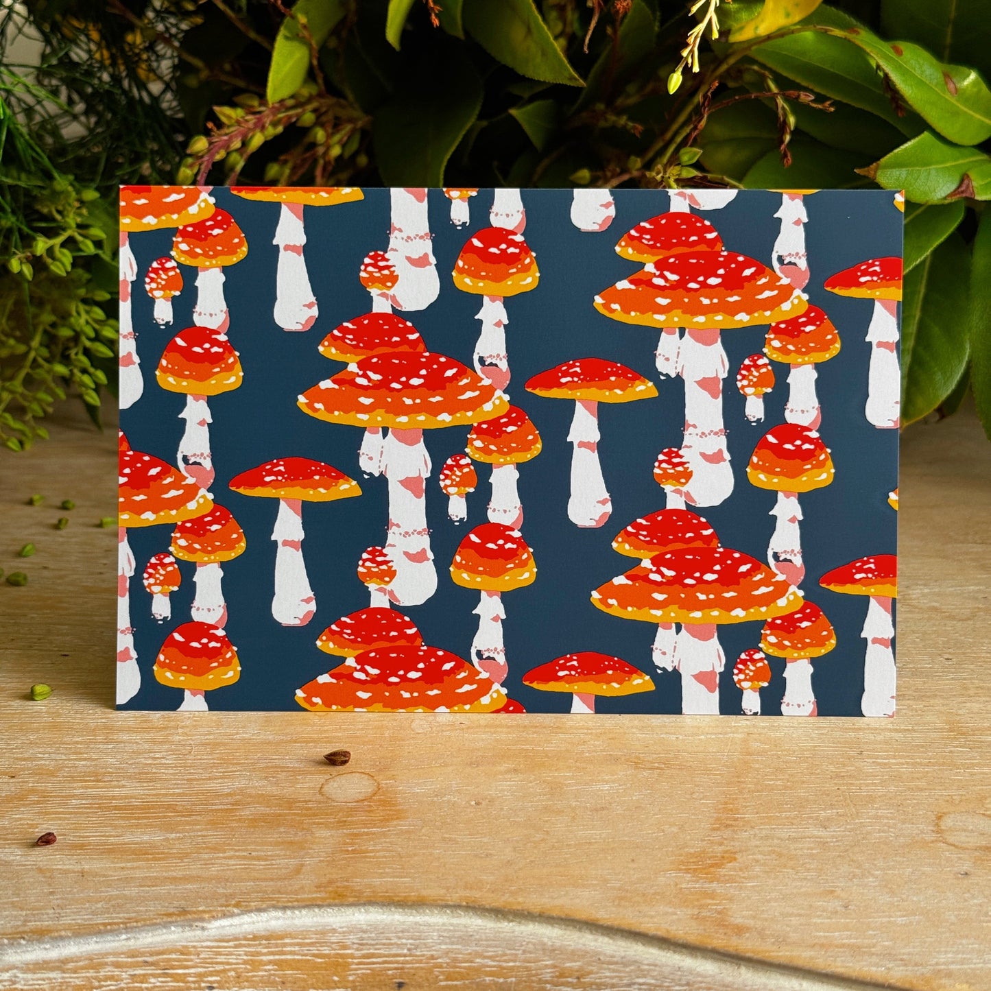 Card with red and white mushroom pattern on a blue background, placed on a wooden surface with green leaves in the background.