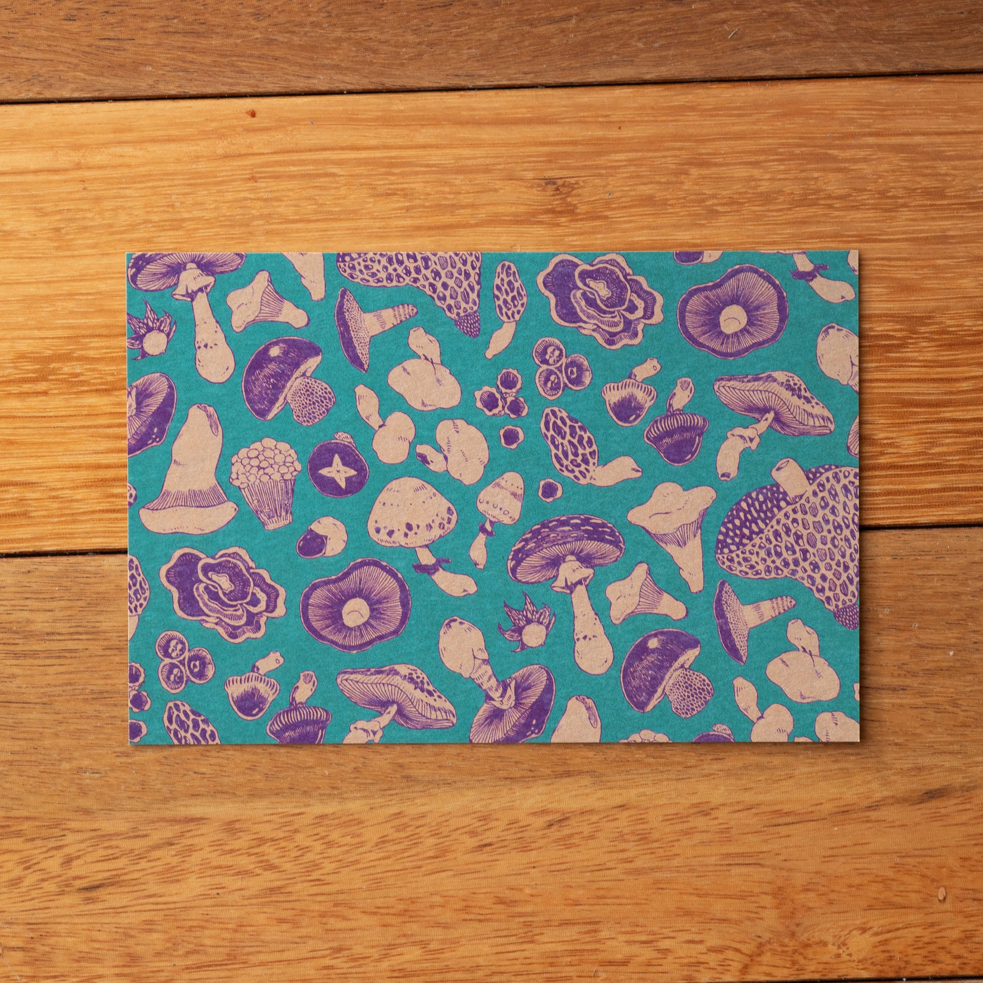 Patterned mushroom postcard with purple and white designs on a teal background, placed on a wooden surface.